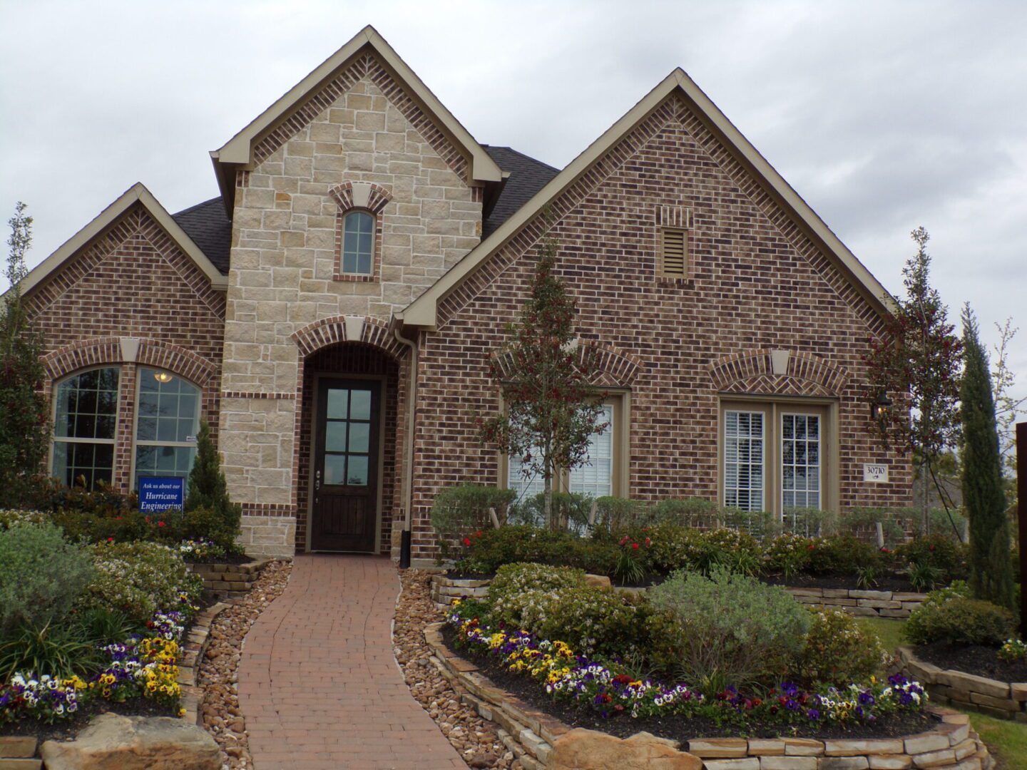 A stone facade two-story house with a landscaped garden and a brick pathway leading to a wooden front door, under a cloudy sky, constructed by Texas builders.