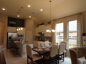 A modern kitchen and dining room designed by Texas builders, featuring white cabinetry, pendant lights, and a dining table set for six, adorned with a "gather" sign on the wall.