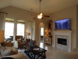 Spacious living room with neutral decor, featuring a fireplace, large windows with curtains, a ceiling fan, and a wall-mounted TV displaying a nature scene crafted by Texas builders.