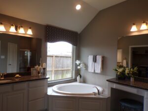 Modern bathroom with a large corner bathtub, gray cabinets, granite countertops by Texas builders, and a small window with patterned curtains.