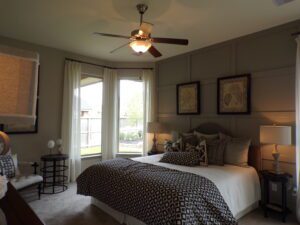 A well-appointed bedroom featuring a large bed with patterned bedding, two side lamps, windows with beige curtains, and a ceiling fan. Decor includes framed wall art and a neutral color scheme by Texas builders