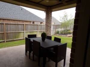 View of a patio with a black wicker dining set under a covered area, built by Texas builders, overlooking a grassy backyard enclosed by a wooden fence.