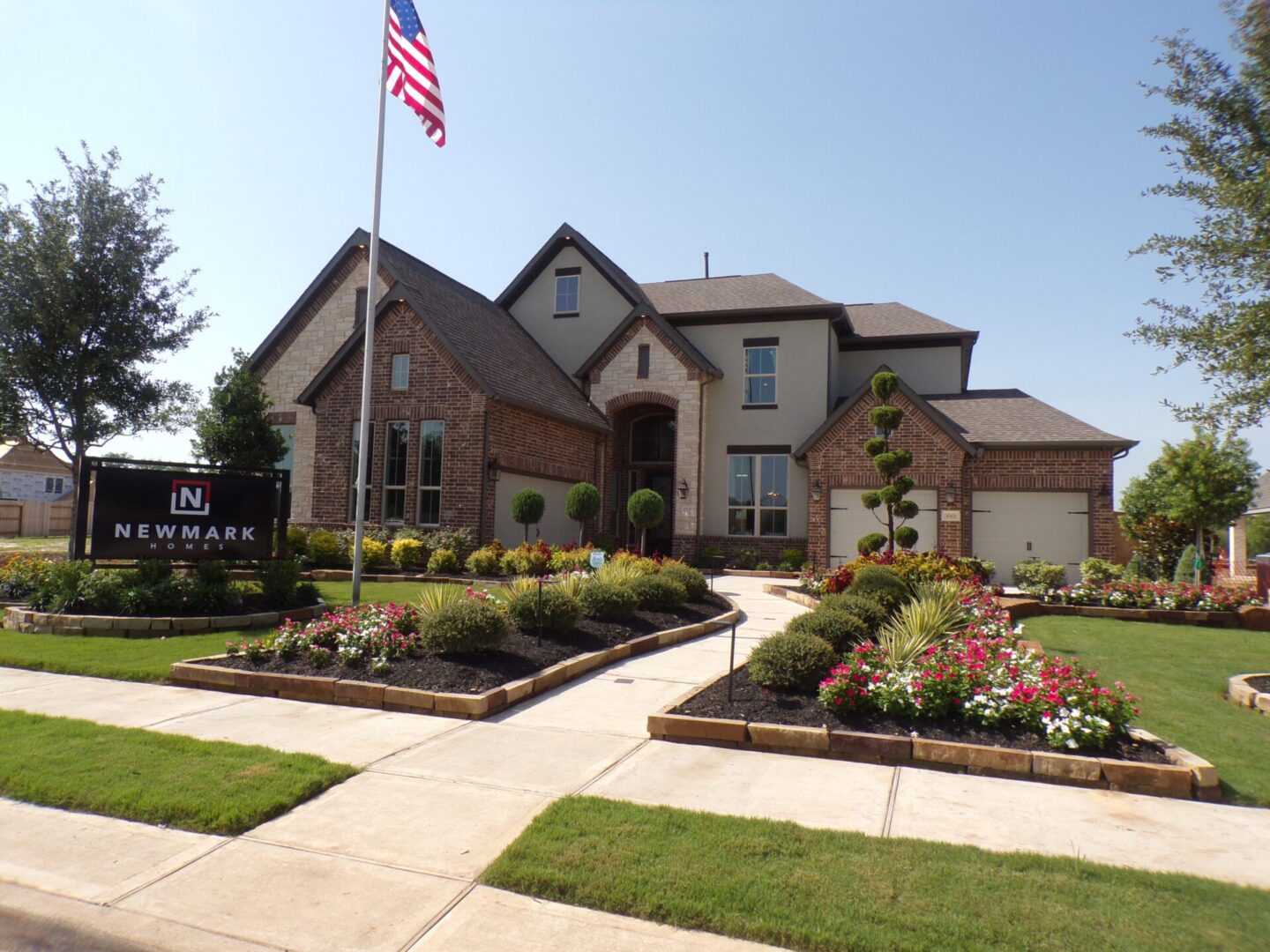 A large two-story brick house with a landscaped front yard and an American flag, under a clear sky, crafted by Texas builders.
