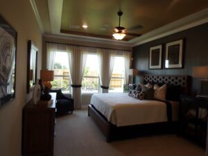Elegant bedroom with a large bed, side tables, lamps, wall art, and bay windows crafted by Texas builders, offering views of an outdoor patio.