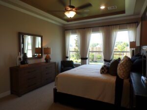 A well-lit bedroom with a large bed, wooden dresser, mirror, and window curtains crafted by Texas builders overlooking a garden.