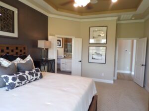 Elegant bedroom featuring a large bed with decorative pillows, framed art on walls, tray ceiling, and open doors leading to a closet and bathroom, designed by Texas builders.