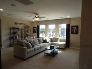 Spacious living room with a large beige sofa, coffee table, ceiling fan, and a shelving unit displaying decor by Texas builders. Large windows provide abundant natural light.