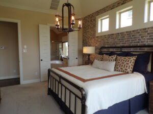 A well-appointed bedroom featuring a king-size bed with navy and white bedding, an exposed brick wall constructed by Texas builders, rectangular pendulum light fixtures, and doors leading to an en-suite bathroom.