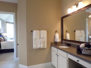 A neutrally decorated bathroom crafted by Texas builders, featuring a large mirror, double sink vanity, white towels, and a view into a bedroom.