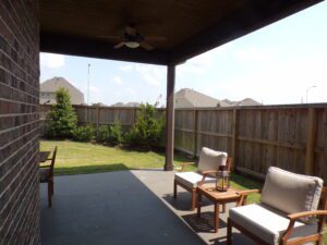 Covered patio with two chairs facing a wooden fence, ceiling fan above, and a sunny Texas suburban landscape visible beyond the fence.