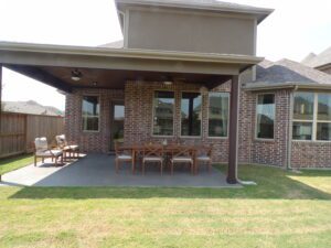 An outdoor patio area with a dining table and a seating arrangement under a covered section next to a brick house, constructed by Texas builders.