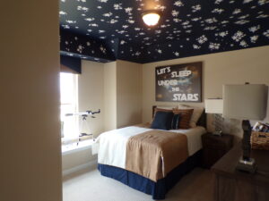 A cozy bedroom with a star-patterned ceiling, a bed with beige and blue bedding, a telescope near a window, and a wall poster that reads "let's sleep under the stars," designed by