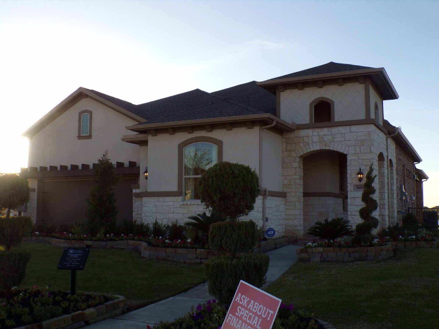 A two-story suburban home with a stone facade and a two-car garage, constructed by Texas builders, surrounded by a manicured lawn and promotional real estate signs.