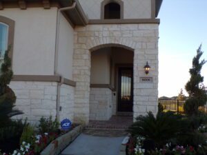 Front entrance of a modern house with a stone archway, adorned with flowers, leading to a wooden door marked "8006", constructed by Texas builders.