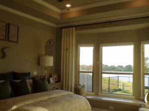 A cozy bedroom with a bed, side lamp, and large windows overlooking a lake, decorated in earth tones and featuring framed art by Texas builders on the walls.