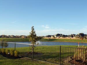 A suburban landscape featuring a fenced pond, a freshly planted tree, and a row of houses built by Texas builders under a clear sky.