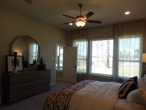 A well-lit bedroom with a large bed, dresser, mirror, ceiling fan, and windows with blinds overlooking trees, crafted by Texas builders.