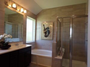 Modern bathroom with beige tiled walls, a glass-walled shower, a bathtub, dark wooden vanity by Texas builders, and an orchid beside an art piece.