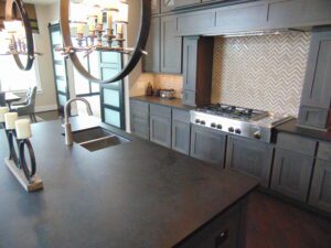 Modern kitchen with dark gray cabinets, chevron backsplash, a gas stove, and an island with a sink and decorative candles, crafted by Texas builders.