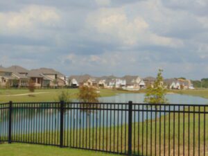 View of a suburban neighborhood featuring modern homes along a lake, bordered by a black iron fence under a partly cloudy sky, constructed by Texas builders.