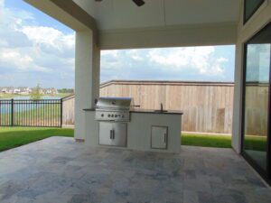 A covered patio featuring a built-in barbecue grill with a stone countertop, constructed by Texas builders, overlooking a fenced backyard under a cloudy sky.