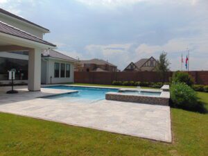 A suburban backyard featuring a rectangular swimming pool with a spa, built by Texas builders, surrounded by a landscaped lawn and a modern house on a sunny day.