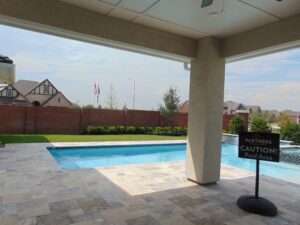 Covered patio overlooking a rectangular pool with a caution sign next to it, set against a backdrop of Texas builders' residential houses and a clear sky.
