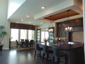 Modern kitchen with dark cabinetry, bar stools, and double chandelier, extending to a dining area with a large table under a coffered ceiling designed by Texas builders.