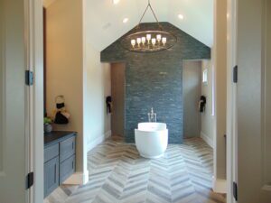 A modern bathroom featuring a freestanding tub, gray herringbone tile floor, teal stone accent wall, and an elegant overhead chandelier constructed by Texas builders.
