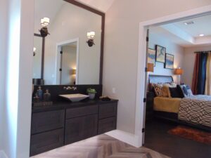 A modern bathroom with a large mirror and dark wood vanity, designed by Texas builders, connects to a bedroom with an ornate bedspread.