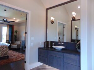 Interior view of a bathroom with dark wood cabinets, a white countertop basin, and a mirror, designed by Texas builders, leading to a visible bedroom through an open door.