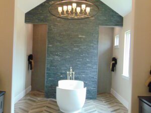 Modern bathroom with herringbone tiled floors, a standalone white bathtub in front of a dark stone accent wall, illuminated by an overhead chandelier crafted by Texas builders.