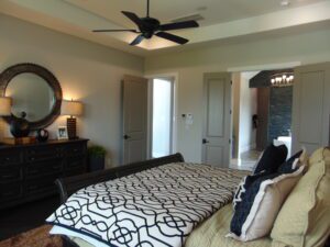 Modern bedroom featuring a bed with geometric-patterned bedding, a dark wood dresser with a round mirror, a ceiling fan installed by Texas builders, and an open door leading to another room.