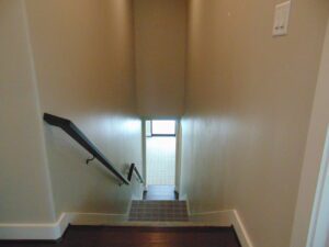 Interior view of a stairway leading down to a landing with a window, featuring handrails on the left and light neutral walls constructed by Texas builders.