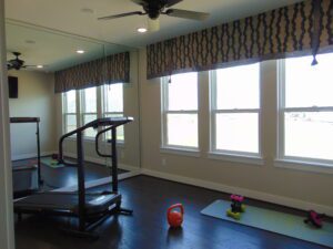 Home gym with a treadmill, yoga mats, weights, large windows with blinds crafted by Texas builders, and patterned valances. Ceiling fans and recessed lighting overhead.
