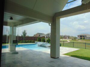 View from a covered patio of a backyard with a pool and suburban houses built by Texas builders in the distance under a cloudy sky.