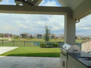 View of a suburban landscape with houses and a lake from a patio featuring a ceiling fan and a grill, constructed by Texas builders.