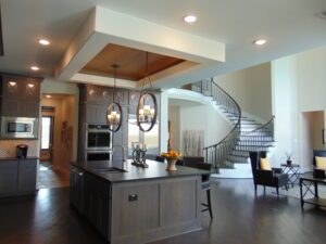 Modern kitchen interior with a central island, pendant lights, and a spiral staircase in the background. Bright and spacious room with wooden and grey cabinets, crafted by Texas builders.