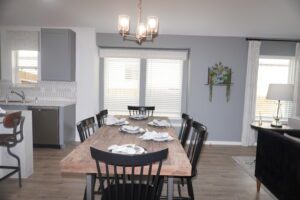 Modern dining room with a wooden table set for six, gray chairs, pendant lights, and an adjacent kitchen area designed by Texas builders.