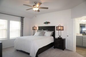 A neatly arranged bedroom constructed by Texas builders with a black bed frame, white bedding, wooden nightstands with lamps, and a ceiling fan above, showcasing a clean, modern decor.