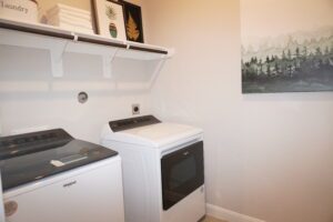 A laundry room with a white washer and dryer, shelves with towels and decorations, and a large painting of a forest on the wall, designed by Texas builders.
