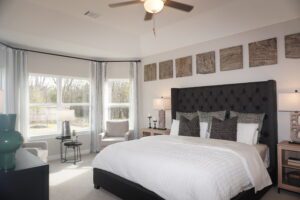 Modern bedroom featuring a large bed with dark headboard, white bedding, two nightstands, and a large window with curtains. Decor includes wall art and a ceiling fan designed by Texas builders.