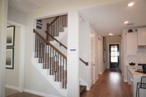 Modern house interior featuring a staircase with wooden handrail and black metal balusters, a hallway leading to a kitchen, and white walls, constructed by Texas builders.
