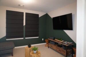 Modern living room with dark green accent walls, three covered windows, a wall-mounted TV, and stylish wooden furniture designed by Texas builders.