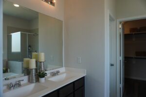 A modern bathroom vanity with a large mirror reflecting a shower area, alongside decorative candles, adjacent to an open door leading by Texas builders to a closet.