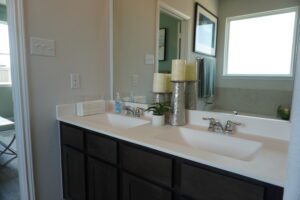 A modern bathroom with a double sink vanity, large mirror, and decorative items on the countertop. Two windows installed by Texas builders provide natural light.