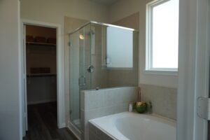 Modern bathroom with glass shower, bathtub, and adjoining walk-in closet, featuring tiled walls and floors with a bright window, constructed by Texas builders.