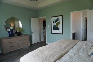 A neatly arranged bedroom with teal walls, wooden flooring, a bed with a cream quilt, a round mirror, a framed abstract painting by Texas builders, and two white doors.