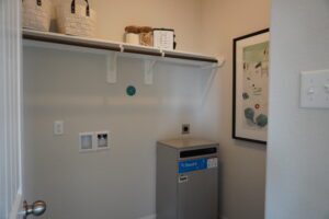 A small room featuring a white shelf with baskets, a wall-mounted framed abstract art piece by Texas builders, and a silver shredder near light switches.