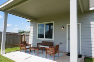 A covered patio area with a white door and two wooden benches on a concrete slab, adjacent to a lawn and enclosed by a Texas builders' fence under a clear sky.